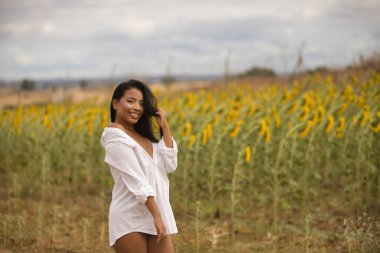 Genç, güzel, Latin esmer kadın beyaz gömlek giyiyor, tatlı ve narin, ayçiçeği tarlasının yanında poz veriyor. Kavram: doğa, tarım, kırsal.