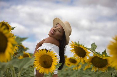 Beyaz bir silindir ve şapka giyen güzel Latin esmer bir kadının portresi, ayçiçeği tarlasında bitkilerin arasında kendini kucaklıyor ve seviyor. Kavram: doğa, tarım, kırsal.