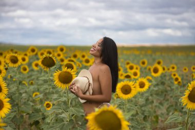 Genç, güzel, Latin esmer kadın beyaz bir bikini giyiyor ve göğsünü şapkayla örtüyor. Ayçiçeği tarlasının ortasında şehvetli ve kışkırtıcı bir şekilde poz veriyor. Kavram: Doğa.