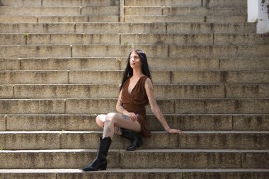 Genç bir kadın, esmer, uzun boylu, zayıf ve çok dövmeli, kahverengi elbiseli ve çizmeli, tatlı, masum ve yalnız, buharlaşmış, merdivenlerde oturuyor. Gençlik kavramı, güzellik, asilik, milenyum.