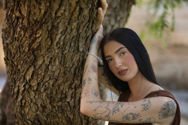 Genç bir kadının portresi, esmer, uzun boylu, ince ve çok dövmeli, kahverengi bir elbise giyiyor, kameraya tatlı, şefkatli ve masum bir şekilde bakıyor, bir ağacın gövdesine yaslanıyor..