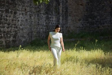 Beyaz elbiseli, genç, güzel, esmer bir kadın, rahat ve sakin, güneşli bir bahar gününde uzun çimlerde yürüyor. Güzellik, moda, moda, gençlik kavramı.