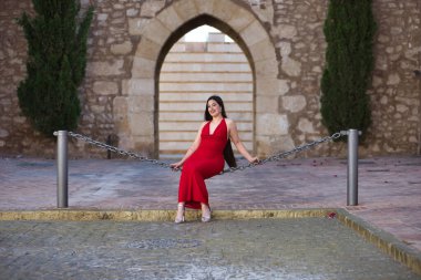 Genç, güzel, esmer, güzel kırmızı parti elbisesi içinde gülümseyen, mutlu ve komik poz veren bir kadın sokakta büyük bir zincirde oturuyor. Güzellik, moda, moda, gençlik kavramı.