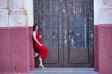 Genç, güzel, esmer, güzel bir kadın güzel kırmızı bir parti elbisesi ve topuklu ayakkabıları içinde, eski bir kapının çerçevesine yaslanmış. Güzellik, moda, moda, gençlik kavramı.