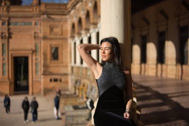 Genç, güzel, esmer kadın portresi, siyah süslü bir elbise giyiyor, gözleri kapalı, güneş ışınlarını alıyor, mermer bir sütuna yaslanıyor ve oturuyor. Güzellik kavramı, moda, kadınlık.