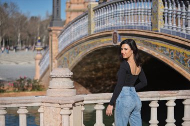 Genç, güzel, siyah uzun kollu tişörtlü ve kot pantolonlu bir kadın, arkası dönük, kameraya bakıyor, Seville 'de İspanya meydanında bir korkuluğa yaslanıyor..