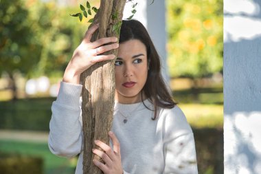 Beyaz elbiseli, genç, güzel, koyu renk saçlı bir kadının portresi, tatlı ve rüya gibi bakışlı, bir sarmaşığın gövdesine yapışan. Güzellik, moda, eğilim, kadınlık, doğa, bitkiler, ağaçlar.