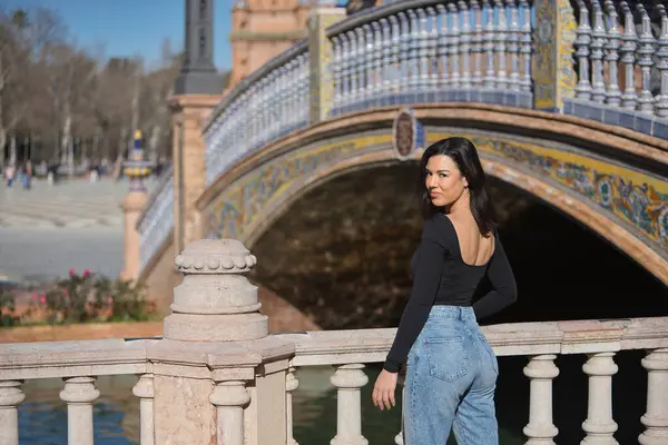 Genç, güzel, siyah uzun kollu tişörtlü ve kot pantolonlu bir kadın, arkası dönük, kameraya bakıyor, Seville 'de İspanya meydanında bir korkuluğa yaslanıyor..