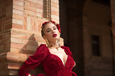 Genç, güzel, sarışın bir kadının portresi, kırmızı flamenko elbisesi, saçında çiçekler, altın mücevherler, flamenko pozları, Seville 'de İspanya meydanında kameraya bakarken.