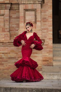 Genç, güzel, sarışın, kırmızı flamenko elbiseli, saçında çiçekler ve altın mücevherlerle İspanya meydanında flamenko dansı yapan bir kadın. Dans konsepti, sanat. Dikey konum.
