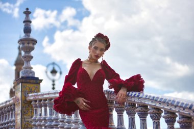 Genç, güzel, kırmızı flamenko elbiseli sarışın kadın, saçında çiçekler ve altın mücevherler, İspanya 'nın Seville meydanındaki güzel bir köprüde flamenko pozları veriyor. Dans konsepti, sanat, tipik.