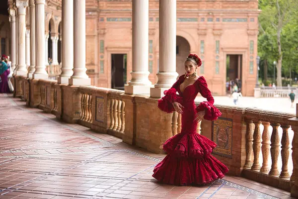 Genç, güzel, sarışın, kırmızı flamenko elbiseli, saçında çiçekler ve altın mücevherlerle İspanya meydanında flamenko pozları veriyor. Dans konsepti, sanat, tipik İspanyol..