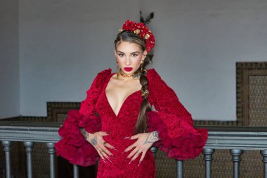 Genç, güzel, sarışın, kırmızı flamenko elbiseli, saçında çiçekler ve altın mücevherlerle, Seville 'de İspanya meydanında metal bir korkuluğa yaslanmış flamenko pozları veriyor. Dans konsepti, sanat.