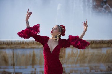 Genç, güzel, sarışın, kırmızı flamenko elbiseli, saçında çiçekler, altın mücevherler, Seville 'de İspanya meydanında fıskiyeli fıskiyelerle flamenko pozları veriyor..