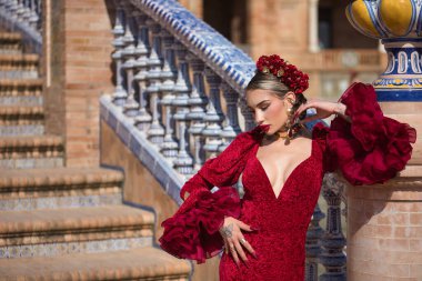 Genç, güzel, sarışın, kırmızı flamenko elbiseli, saçında çiçekler ve altın mücevherlerle, İspanya 'nın Seville meydanında bir tuğla sütuna yaslanmış flamenko pozlar veriyor. Dans konsepti, sanat.