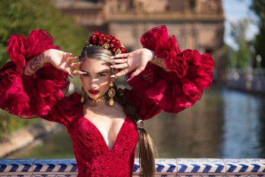 Genç, güzel, sarışın, kırmızı flamenko elbiseli, saçında çiçekler ve altın mücevherlerle, Seville 'de İspanya meydanında fayanslı bir korkuluğa yaslanmış flamenko pozları veriyor. Dans konsepti, sanat.