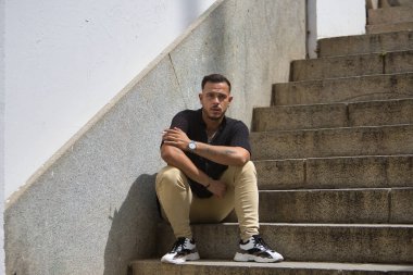 Genç, esmer, İspanyol, siyah tığ işi gömlek ve kot pantolon giyen, sırtı duvara yaslanmış, yalnız bir merdivende oturan modern bir adam. Moda, moda, gündelik konsept.