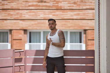 Genç, esmer, İspanyol, beyaz atletli ve kot pantolonlu modern bir adam, ahşap bir çite yaslanmış çapkın bir tavırla poz veriyor. Moda, moda, gündelik konsept.