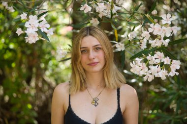Siyah tişörtlü, genç, güzel, sarışın bir kadının portresi, narin ve masum, etrafı beyaz zakkumlarla çevrili, açık hava parkında kameraya bakıyor. Güzelliği kavra, gençliği, milenyumu.