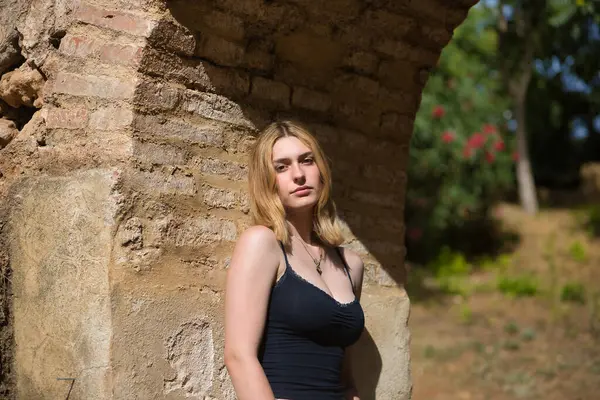 Genç, güzel, sarışın, siyah gömlekli, masum ve hassas bir kadının portresi. Kameraya bakıyor, tuğla kemere yaslanıyor, açık hava parkında güneş ışınları alıyor. Güzellik kavramı, gençlik.