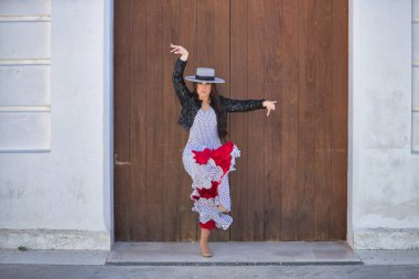 Genç bir kadın, güzel, esmer, flamenko dansçısı, ceketli ve şapkalı güzel bir elbise giyiyor, arka planda ahşap kapılı flamenko dansı yapıyor. Flamenko konsepti, tipik, İspanyolca, Sevilla.