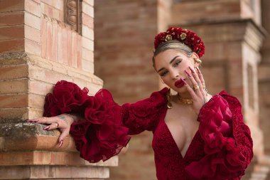 Genç, güzel, kırmızı flamenko elbiseli sarışın kadın, saçında çiçekler ve altın mücevherler, Seville 'de İspanya meydanında tuğla bir duvara yaslanmış poz veriyor. Dans konsepti, sanat, tipik İspanyol..