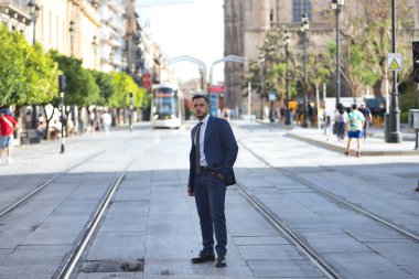 Genç, esmer, İspanyol, takım elbiseli ve kravatlı modern yönetici Seville şehrinde bir caddenin ortasında tek eliyle poz veriyor. Konsept iş, şirket, lider.