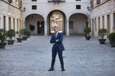 Genç, esmer, İspanyol, modern yönetici, takım elbiseli ve kravatlı, kollarını kavuşturarak poz veriyor, yalnız bir şehir caddesinde başarılı bir adam olduğunu gösteriyor. Konsept iş, şirket, lider.