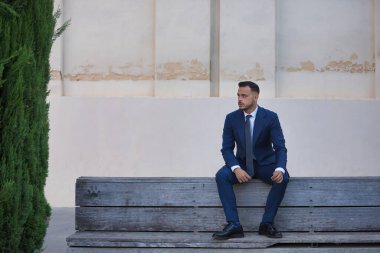 Genç, esmer, İspanyol, modern yönetici, takım elbiseli ve kravatlı, ekonomik kriz konusunda ciddi ve endişeli, mola alıyor, kentteki ahşap bir bankta oturuyor. Konsept iş, şirket, lider.