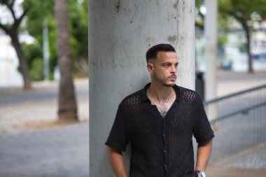 Genç, esmer, İspanyol, siyah tığ işi gömlekli, ciddi ve endişeli bir şekilde poz veren, elleri ceplerinde, bir sütuna yaslanmış modern bir adam. Moda, moda, gündelik konsept.
