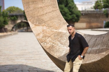 Genç, esmer, İspanyol, modern bir adam, siyah örgü örgü gömlek ve kot pantolon giyiyor, ciddi ve keskin bakışları var, elleri ceplerinde ve taş bir duvara yaslanıyor. Moda, gündelik konsept.