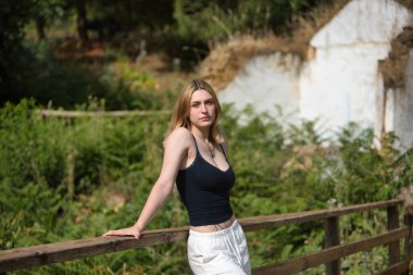 Genç, güzel, siyah tişörtlü, beyaz pantolonlu, yalnız ve melankolik bir kadın, açık hava parkındaki ahşap korkuluklara yaslanmış kameraya bakıyor. Güzelliği kavra, gençliği, milenyumu.