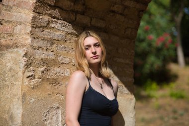 Genç, güzel, sarışın, siyah gömlekli, masum ve hassas bir kadının portresi. Kameraya bakıyor, tuğla kemere yaslanıyor, açık hava parkında güneş ışınları alıyor. Güzellik kavramı, gençlik.