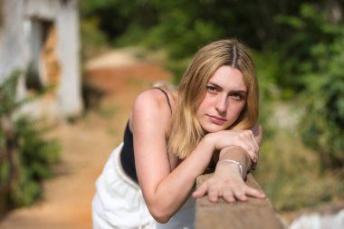 Siyah tişörtlü, beyaz pantolonlu, açık hava parkında tahta korkuluklara yaslanmış genç, güzel sarışın bir kadının portresi. Güzelliği kavra, gençliği, milenyumu.