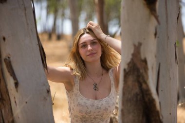 Genç bir kadın, güzel, sarışın, çiçekli bir elbise giymiş, gülümseyen ve mutlu, açık hava parkındaki bir ağacın gövdesine yaslanmış kameraya bakıyor. Güzellik kavramı, gençlik, milenyum.