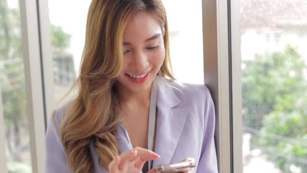 漂亮的亚洲女商人拿着智能手机 与顾客在网上聊天 她站在办公室的窗边 女企业家的概念 — 图库视频影像