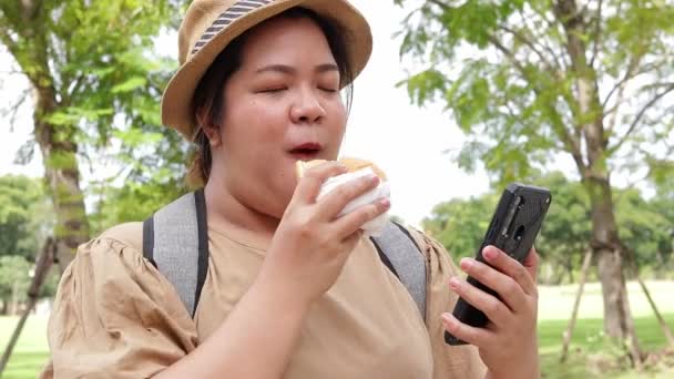 胖女人吃汉堡包 拿着智能手机 一个人旅行的亚洲胖女人快乐生活 旅行概念 — 图库视频影像