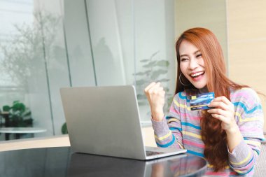 Asyalı bir kadın işte oturuyor ve elinde kredi kartı var. Kolları havada, mutlu, gülümseyen, mutlu. çevrimiçi alışveriş kavramı