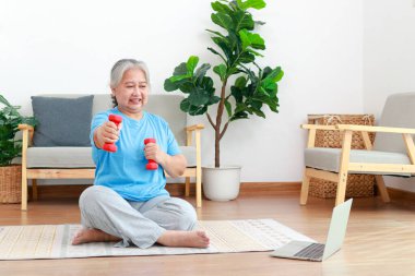 Asyalı son sınıf öğrencisi internetteki antrenörden sonra laptopta egzersiz yapıyor. Yaşlı insanlar evde egzersiz yapmak için dambıl kaldırıyorlar. Spor konsepti. Yaşlılar için sağlık hizmetleri