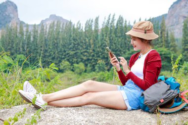 Şapkalı güzel Asyalı kadın gezgin elinde akıllı telefonuyla yerde oturuyor. Neşeli bir şekilde gülümsüyor ve doğa turizminden hoşlanıyor. Seyahat konsepti. Turistler yürüyüş yapar, kamp yapar. Boşluğu kopyala