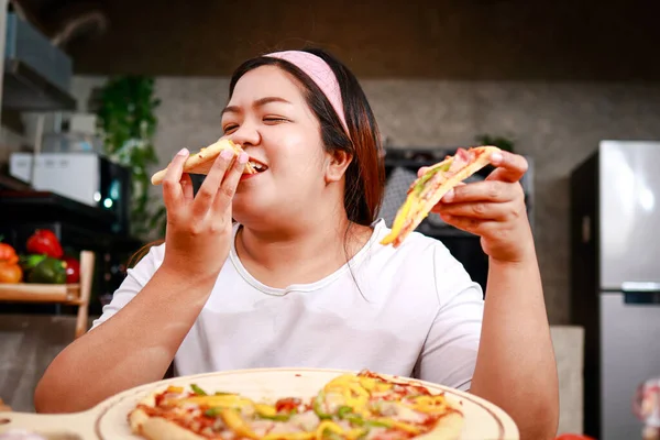 胖胖的亚洲女人吃披萨 她拿着两片披萨 在家里的厨房里美美地吃着 快餐概念 体重减轻 — 图库照片