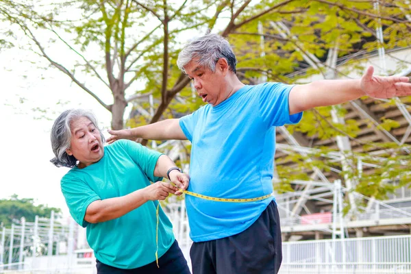 An elderly Asian woman measures the waist circumference of an elderly man with a tape measure. She was shocked by the size of her waist. Sports concept. Health care in retirement. obesity weight loss