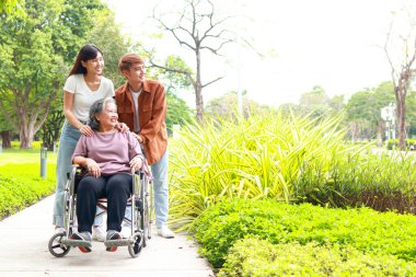 Asyalı aile kavramı. Torun ve torun, büyükanneyi tekerlekli sandalyeyle parkta yürüyüşe çıkarır. Yaşlı sağlık hizmetleri. Boşluğu kopyala