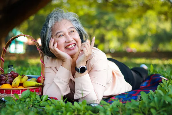Asian senior woman picnicking in the park, smiling, enjoying life. Elderly health care concept