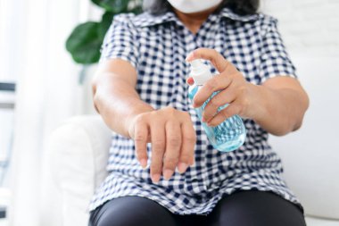 Maske takan yaşlı bir Asyalı kadın virüsü önlemek için ellerine alkol spreyi sıkar. Coronavirus önleme kavramı.
