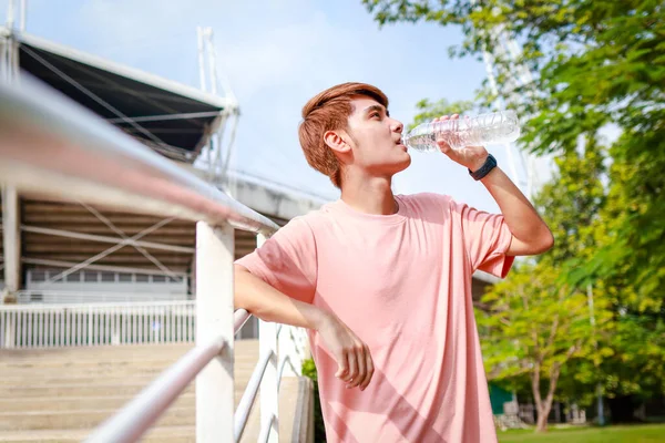 Sabahları dışarıda egzersiz yapan genç Asyalı adam yorgunluğu azaltmak için su içiyor. Spor konsepti. Vücudun sağlık hizmetleri.