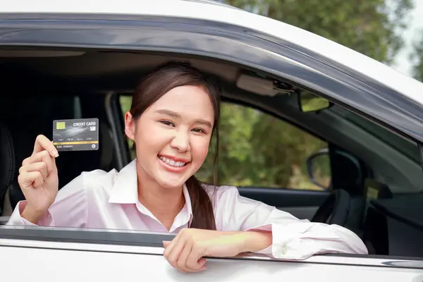 Beautiful Asian woman sitting in a car, happy smile Hold a credit card to pay for car repairs. Car insurance. Card instead of cash. Pay for tires, maintenance costs