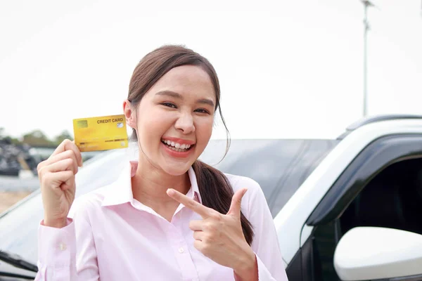 Pretty Asian girl standing next to the car smiling happily. Hold a credit card to pay for car repairs. Car insurance. Card instead of cash. Pay for tires, maintenance costs