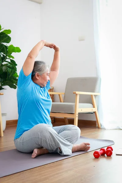 Asian elderly woman exercising at home sitting on the floor in the living room. Sports concept. Elderly health care.