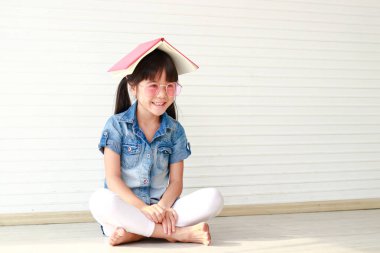 Gözlüklü ve başında kitap olan sevimli bir Asyalı kız. Neşeli ve mutlu bir şekilde gülümsüyor. İlkokul çocuk eğitimi konsepti.